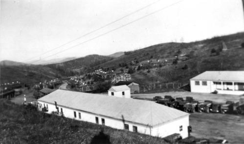 Several large buildings, rows of smaller cabins, and trucks are arranged on the side of a slope. 