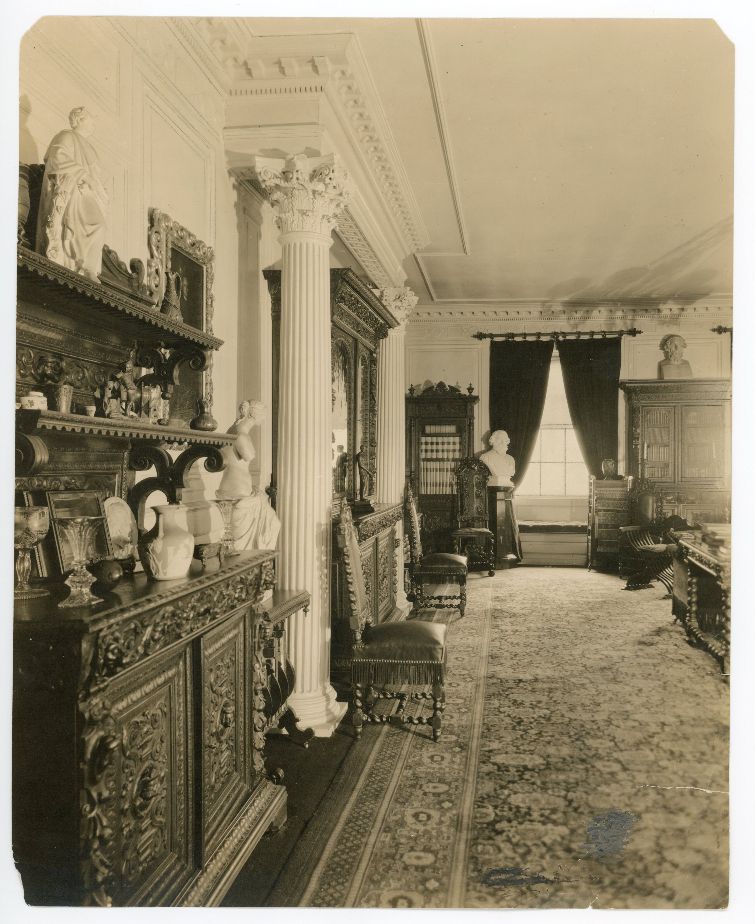 Black and white photograph of 19th century library, with shelves of books, statues, and Greek revival columns featured.