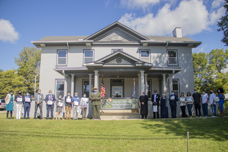 Several people stand in front of a large gray home with two stories with several windows on each level and a peaked roof at the top. The people are lined up next to each other on a concrete sidewalk in front of a green lawn.