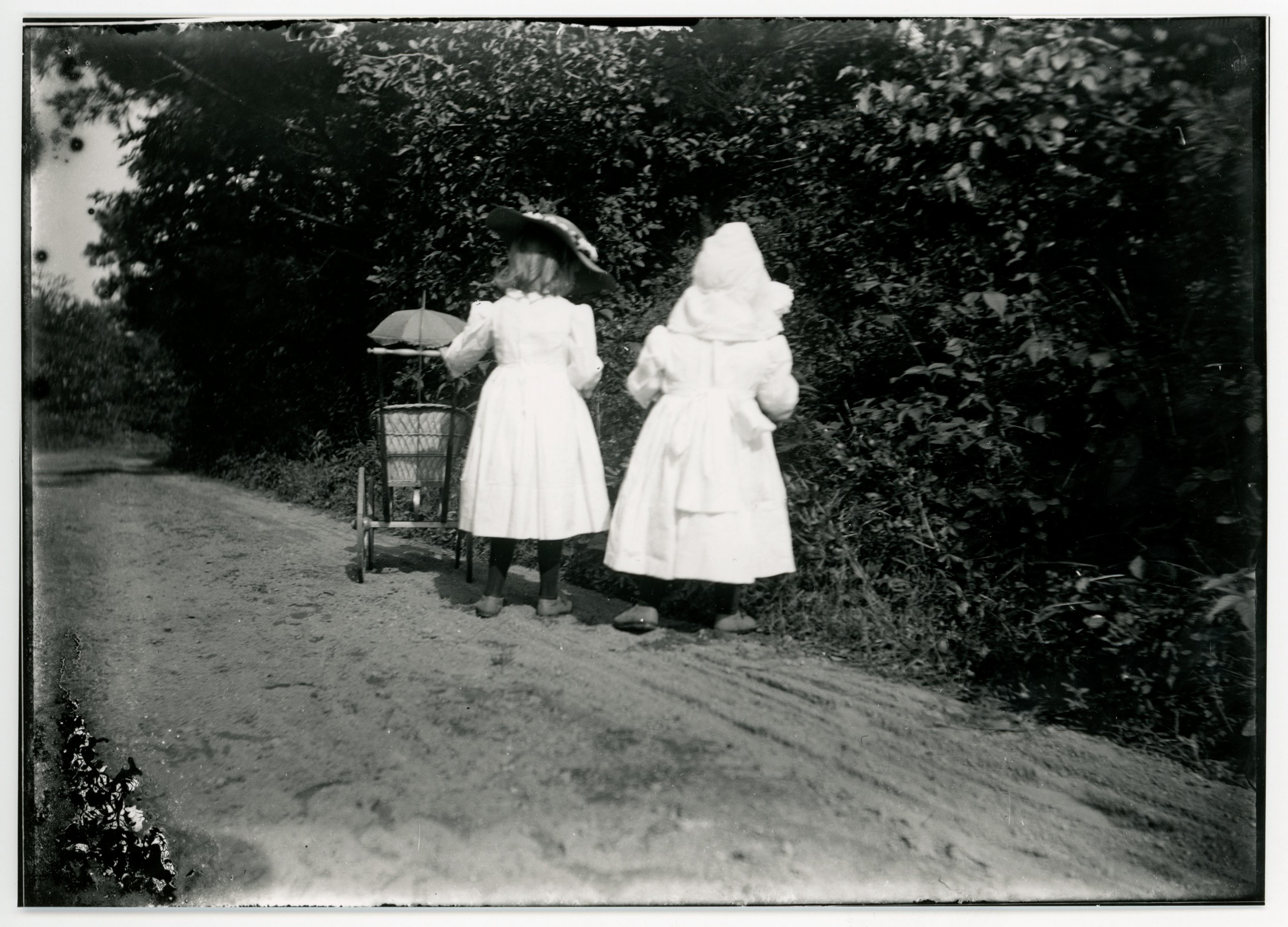 Two young white girls stand on a dirt path looking into large bushes with their backs to the camera. The girl at the left has her left hand on the handle of a wicker doll carriage.