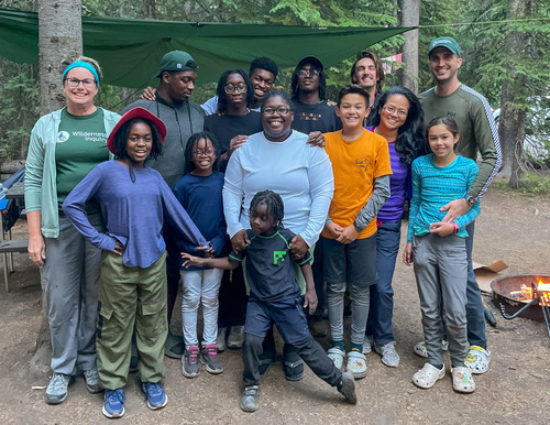 Fourteen adults and children pose for a photo next to a campfire at a campsite.