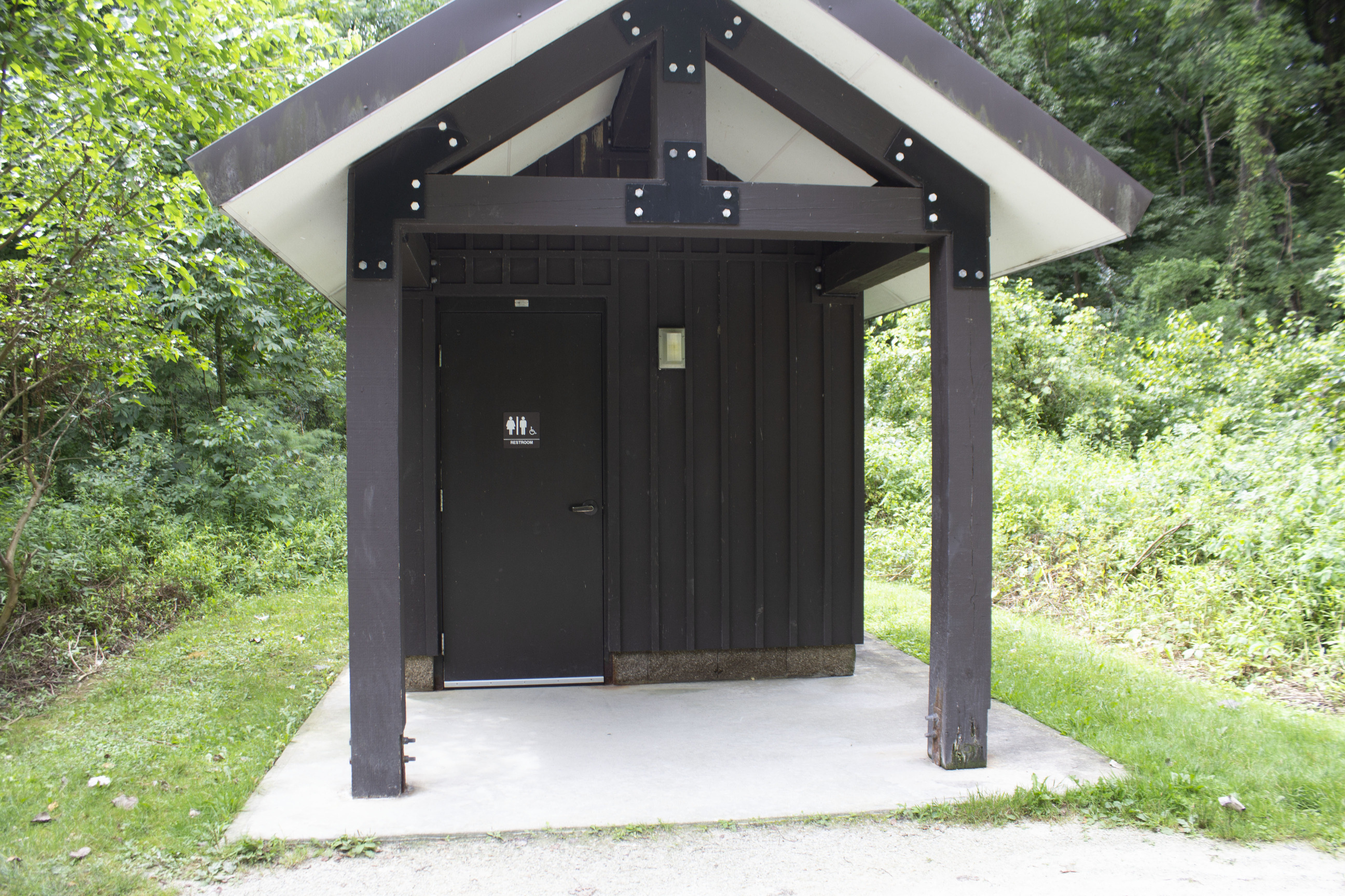 Exterior of brown restroom building with one door, a light, and a unisex sign. An extended roof supported by 2 posts provides shelter.