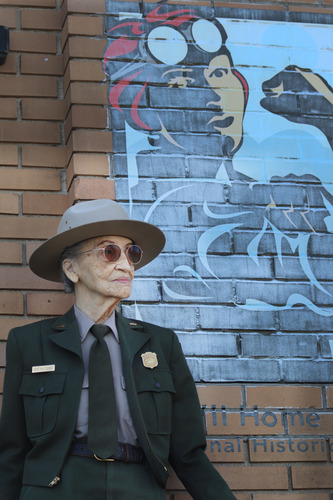 A senior African American woman in a ranger uniform leans against a brick wall. A print of the iconic "We Can Do It" park logo can be see attached to the wall. 