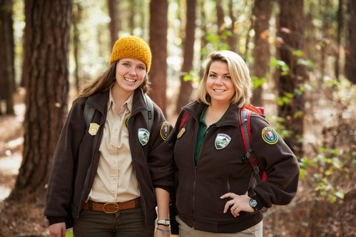 2 young women in brown and khaki uniforms stand and pose for the camera while standing in a sunny forest. 