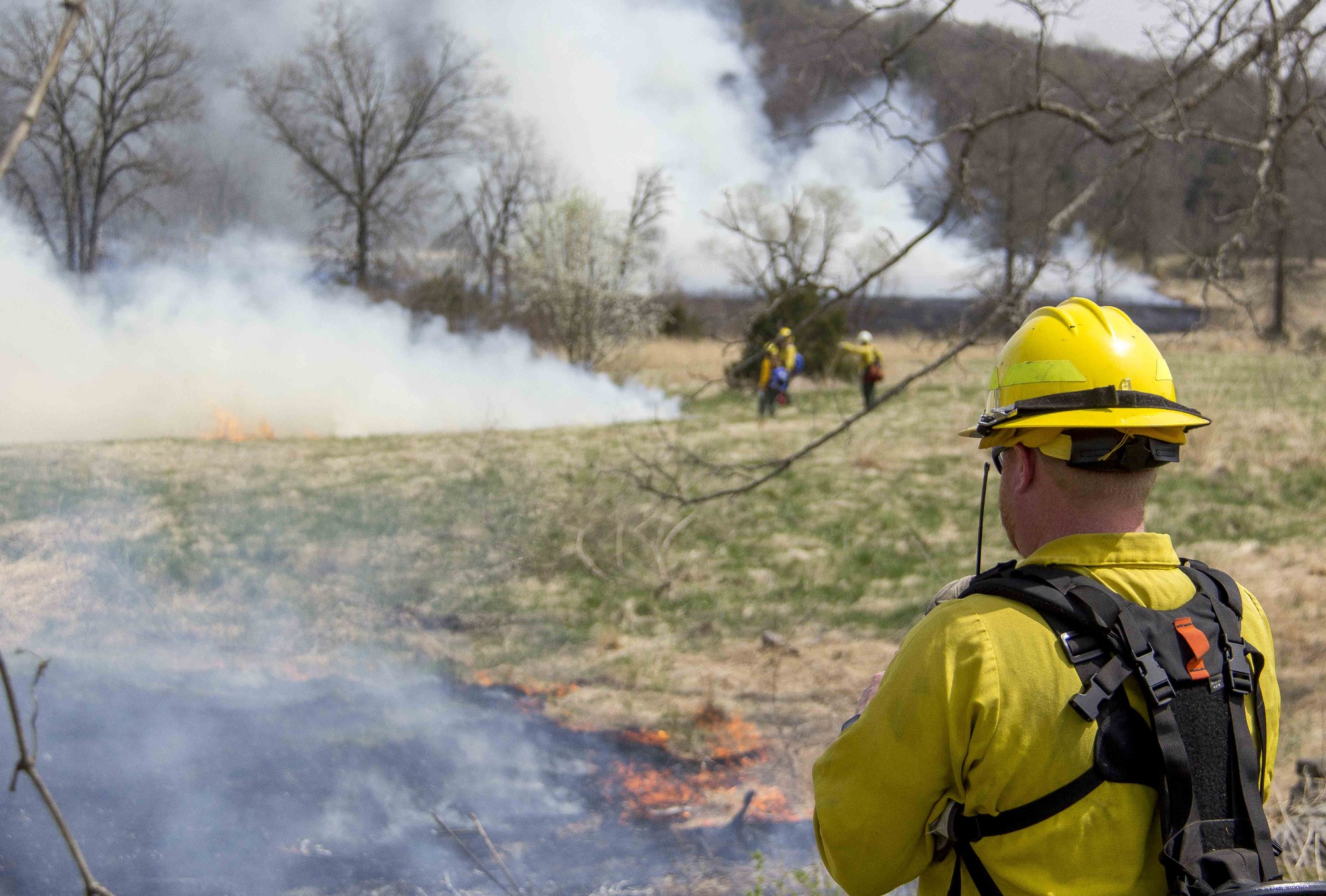 Firefighters monitor the prescribed fire as it moves through the fields south of the Bushman farm. There is a firefighter talking on a hand-held radio in the foreground. He is monitoring the fire. There are firefighters in the background monitoring the fire. There is smoke on the prescribed areas. 