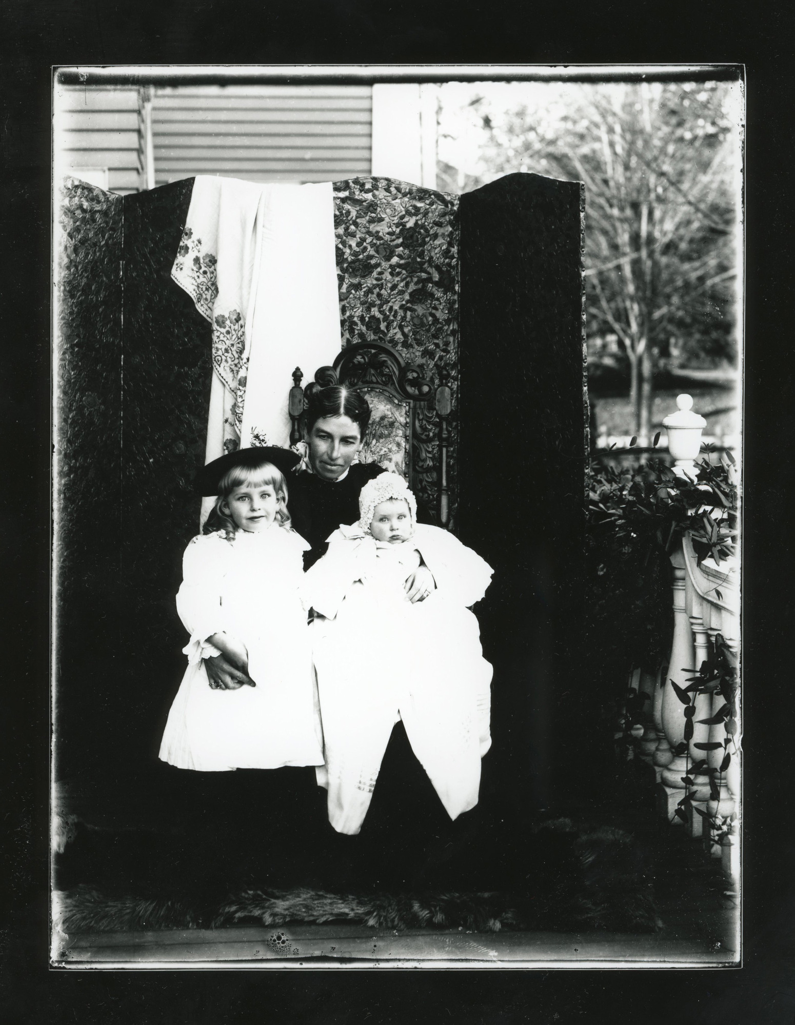 A white woman sits on a chair outdoors holding a baby on her left knee and putting her right arm around a young girl standing next to her. They pose in front of a room divider with a floral design.