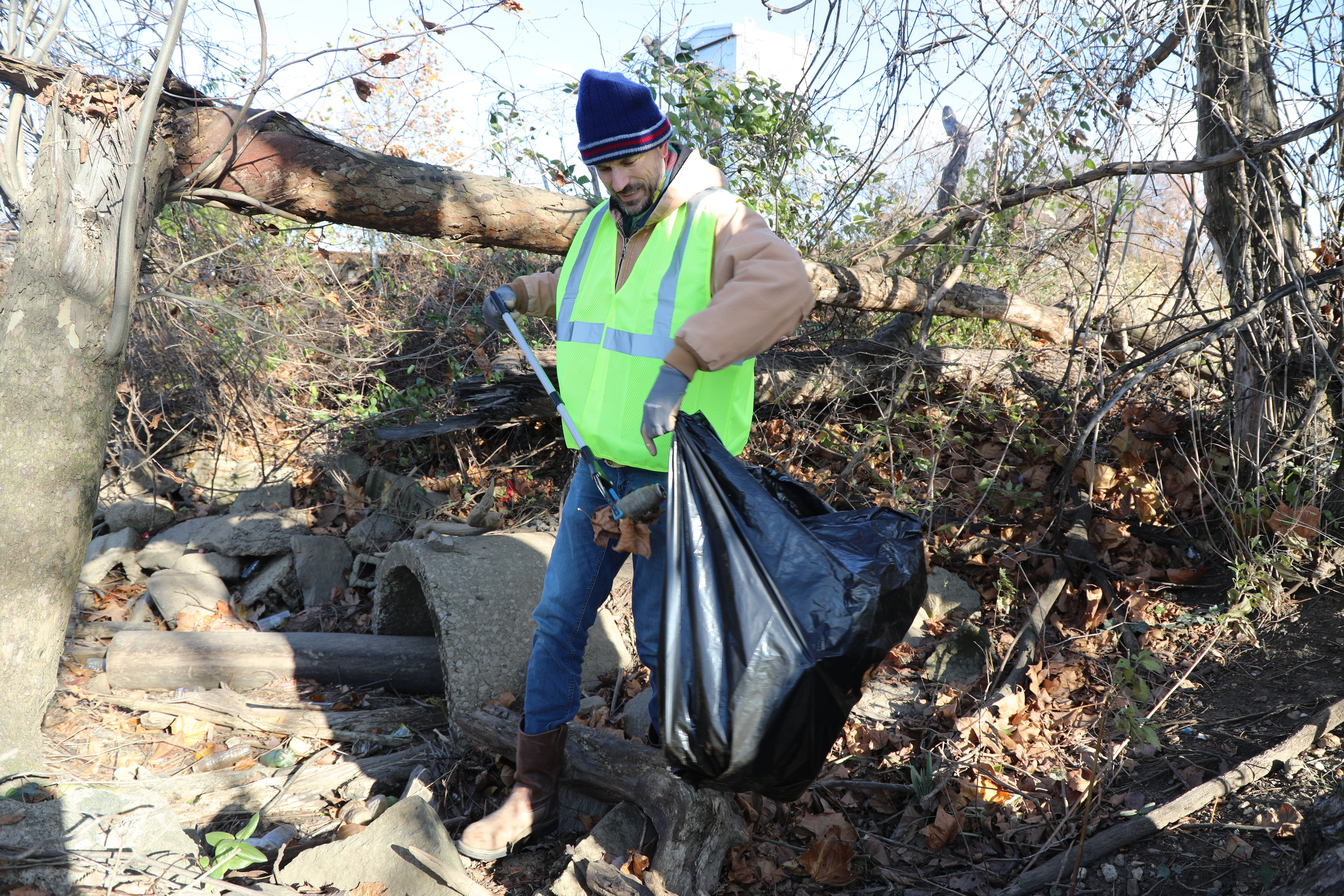 A man wearing a blue knit cap, a light-colored jacket, a bright yellow reflective safety vest, blue jeans, and brown boots is actively cleaning up a debris-strewn area. He is holding a litter picker tool in his right hand and a large black trash bag in his left hand, into which he is placing collected items. The ground around him is covered with leaves, branches, and what appear to be concrete or stone pipes and rubble. In the background, there's a fallen tree trunk and dense vegetation under a clear sky.