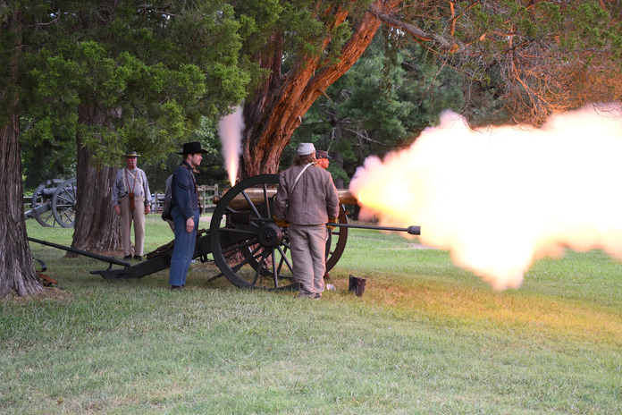 Side view- Five men in confederate uniform fire a cannon with flame and smoke coming out of the vent hole in the back of cannon and the front of the barrel.