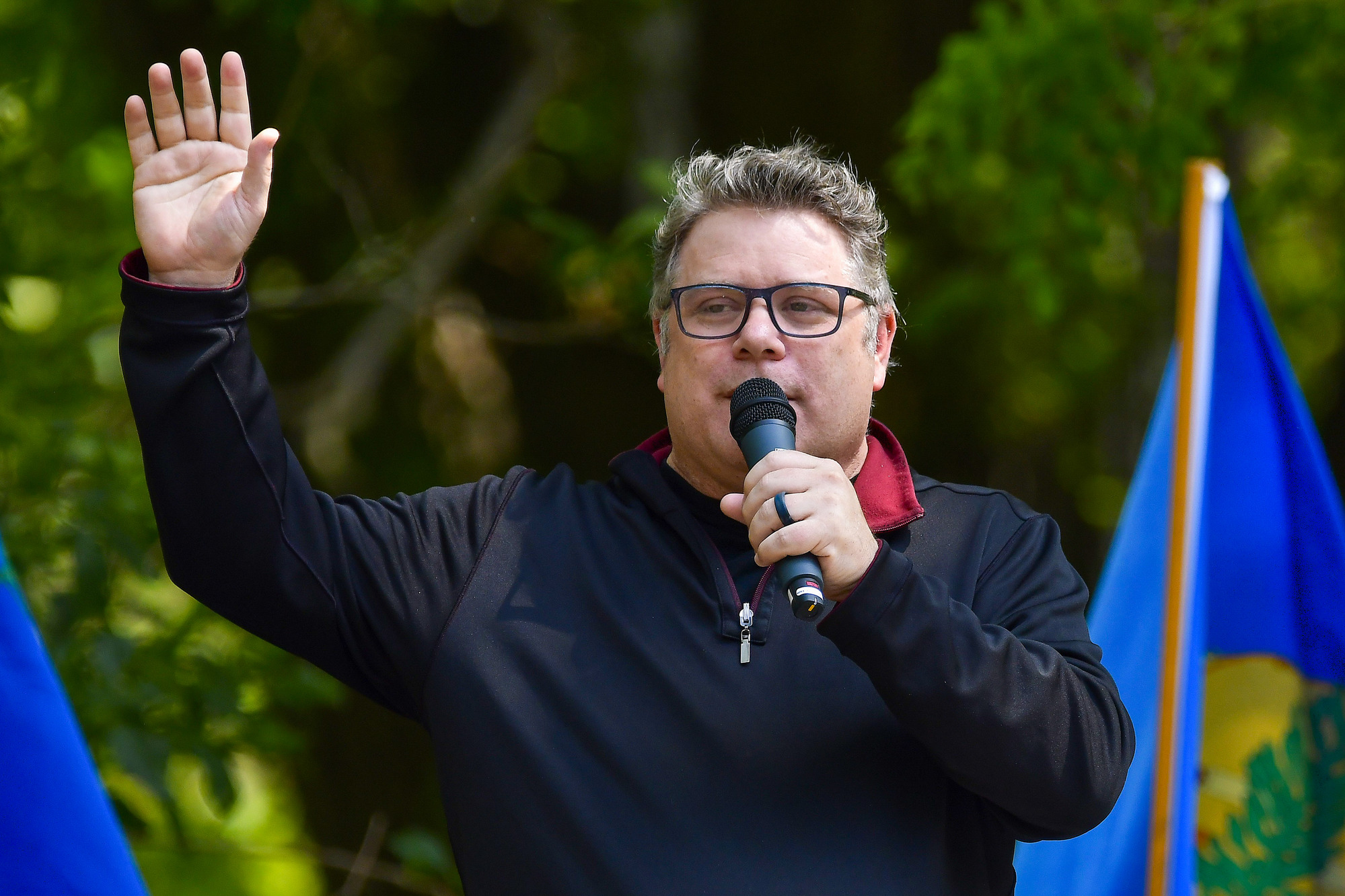 Sean Astin waves and speaks into the microphone during his 2024 Homestead Days keynote speech. He wears glasses and a long sleeve shirt with a red collar and inside lining.