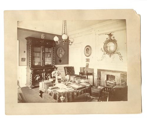 Black and white photograph of 19th century study, circular table in center of room covered in books.