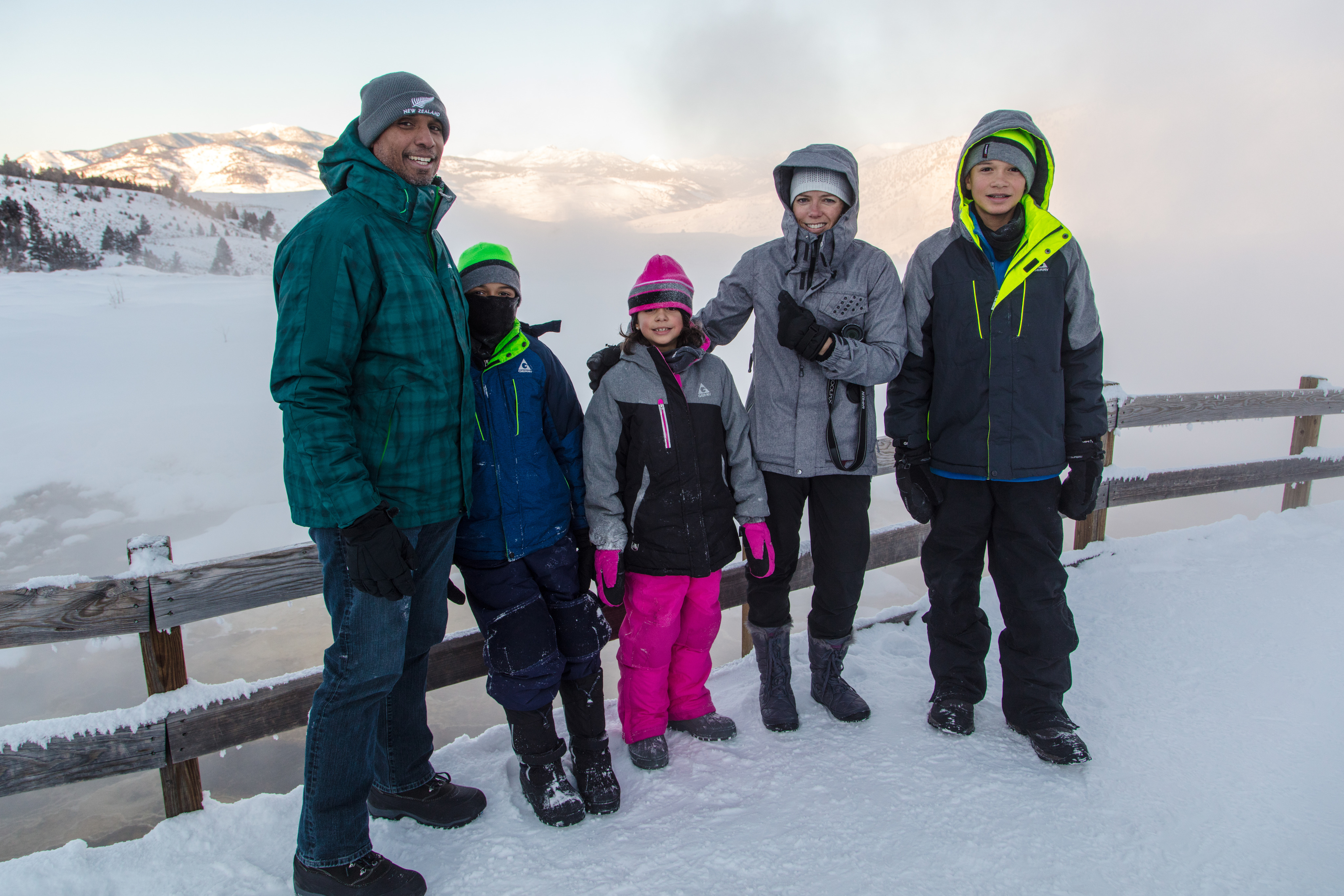 Family of five pose for picture on a snow covered boardwalk in front of a steamy hot spring.