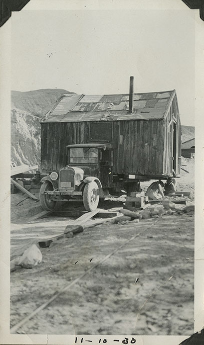 This is an historic black and white photograph from the Scotty's Castle Historic Photograph Collection, Death Valley National Park of Scotty's Castle Scotty's Original Castle on flatbed truck, being set at new location by four laborers. November 10, 1930. Photographed by Mat Roy Thompson.