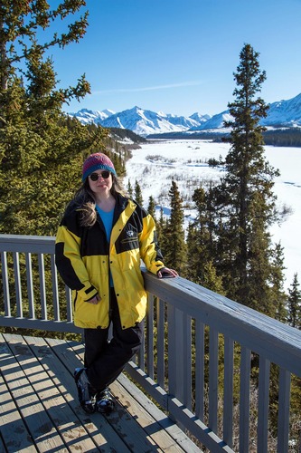 Woman in yellow coat stands by a railing of a deck overlooking a river in winter