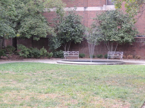 Color photo of fountain and two benches in front of a brick wall lined with trees.