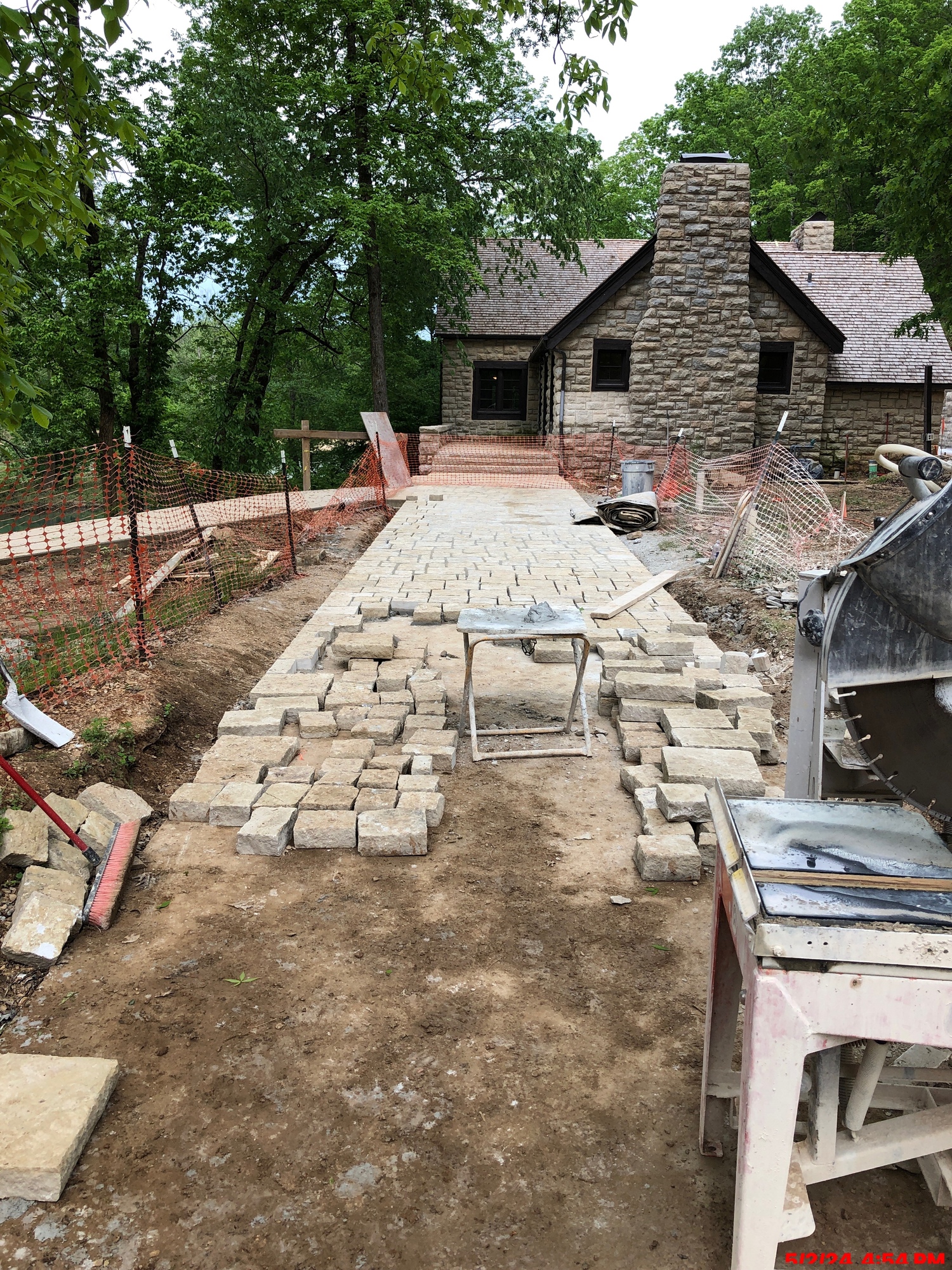 A stone building with a peaked roof and stone chimney on the near side stands among trees in the background with a sidewalk extending towards the viewer. The sidewalk is in the process of being paved with flat flagstones, with work unfinished nearer the viewer.