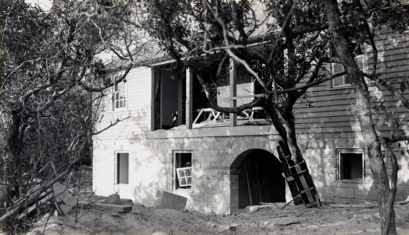 Black and White photo.  Construction debris, lots of sand, windows missing.  Some trees.