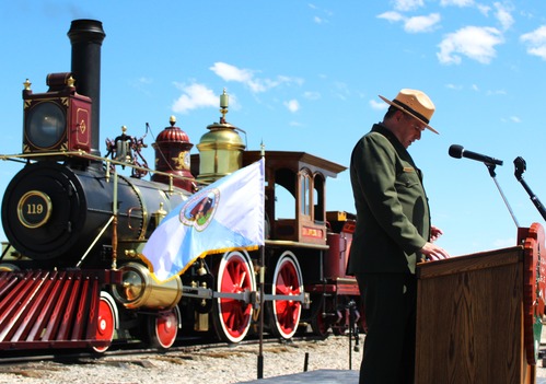 Superintendent Brandon Flint Speaking at podium with 119 Locomotive in background, under blue skies.