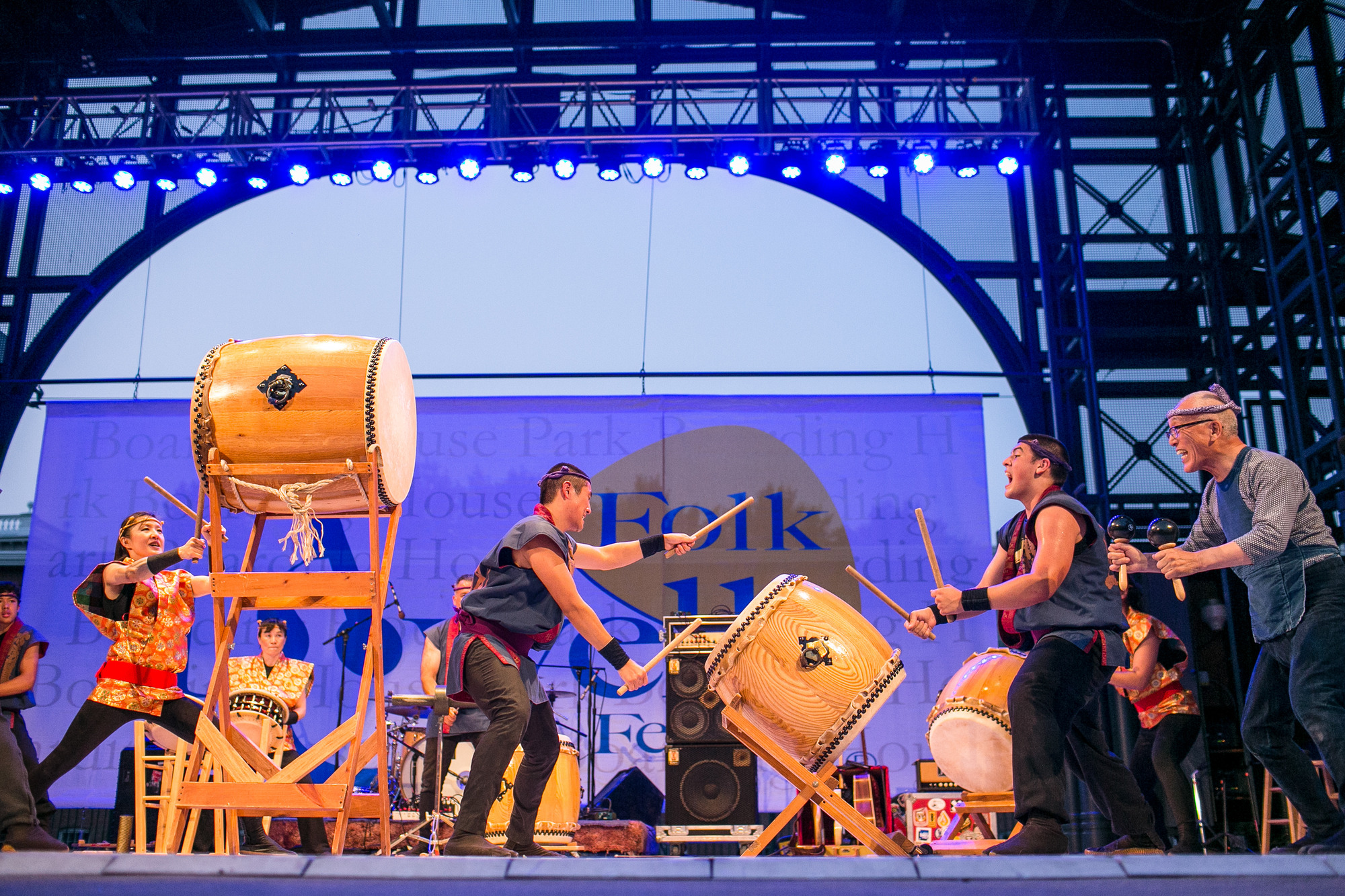 On stage, under the hue of blue lights, performers lean toward the taiko drums with a degree of bend in their knees. Two performers play on a drum on a slanted stand; a performer hits a drum on an elevated stand; and, an older performer has a rattle in each hand.