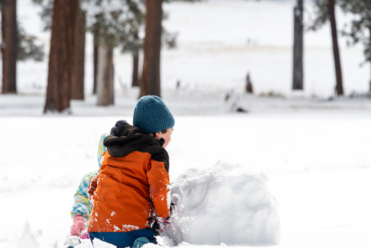Two children kneel in the snow with a giant snowball in front of them.