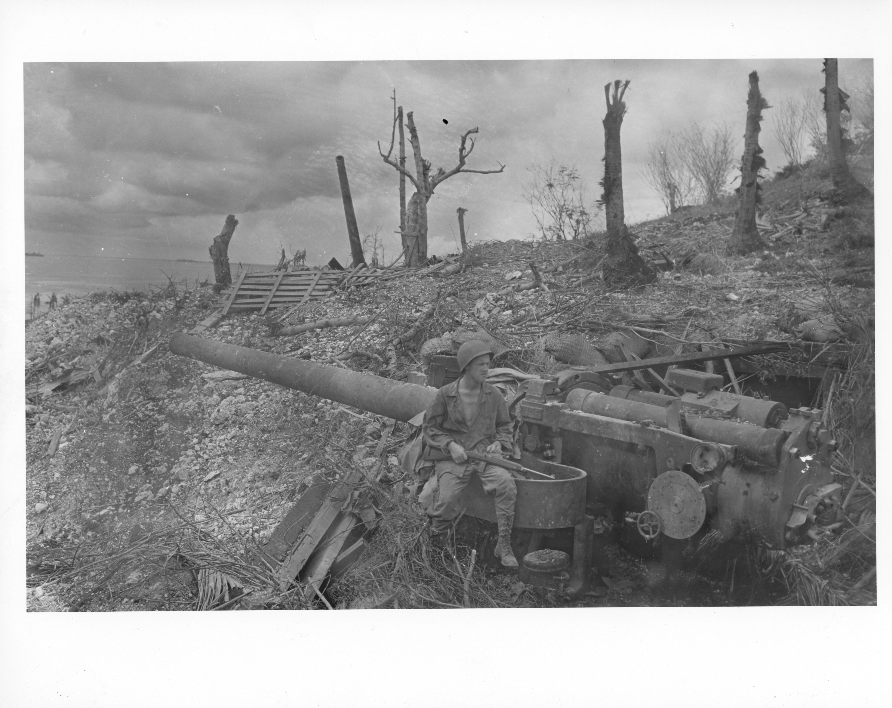 A male soldier in uniform sits on the edge of a large gun. The area around the gun has been badly bombed.