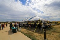 A uniformed ranger is seen to the bottom left speaking with a park volunteer. To their right is a table with green covering where citizenship candidates register their attendance. There is a large blue and white striped tent to the right under which chairs are filling up and a stage can be seen. Blue cloudy sky and green/brown grass.