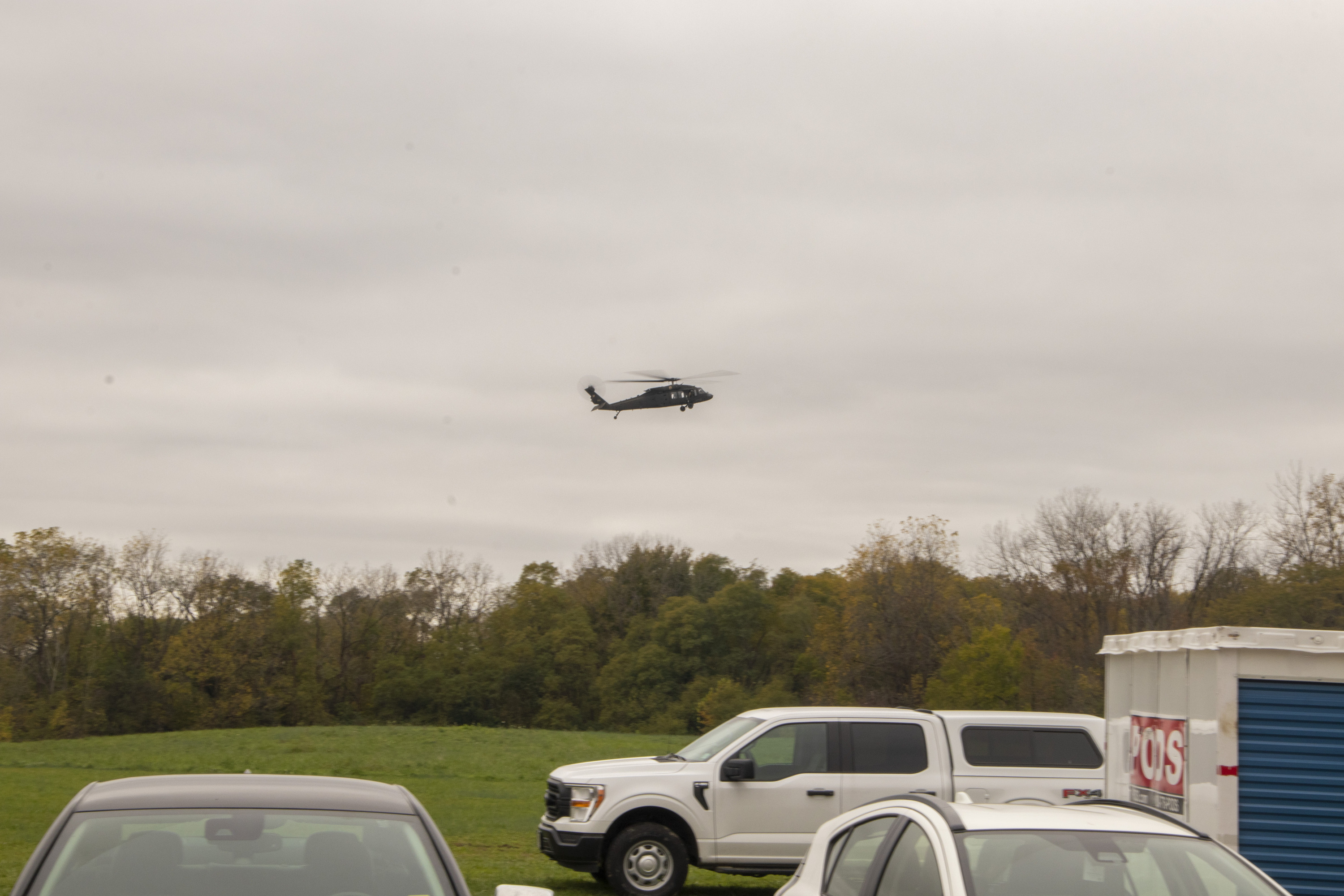 A dark-colored helicopter in flight above multi-colored trees with parked vehicles in the foreground below it.