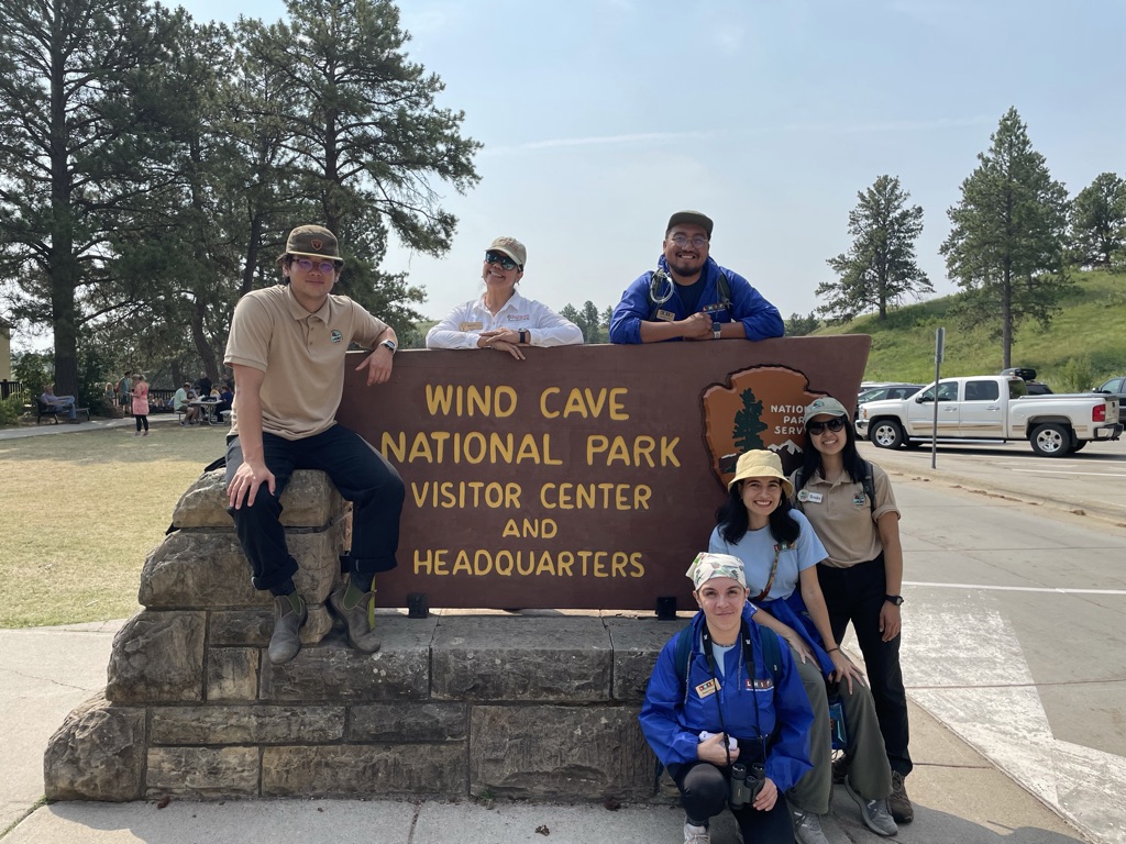 six people smiling and posing with a sign that reads 'wind cave national park visitor center and headquarters'