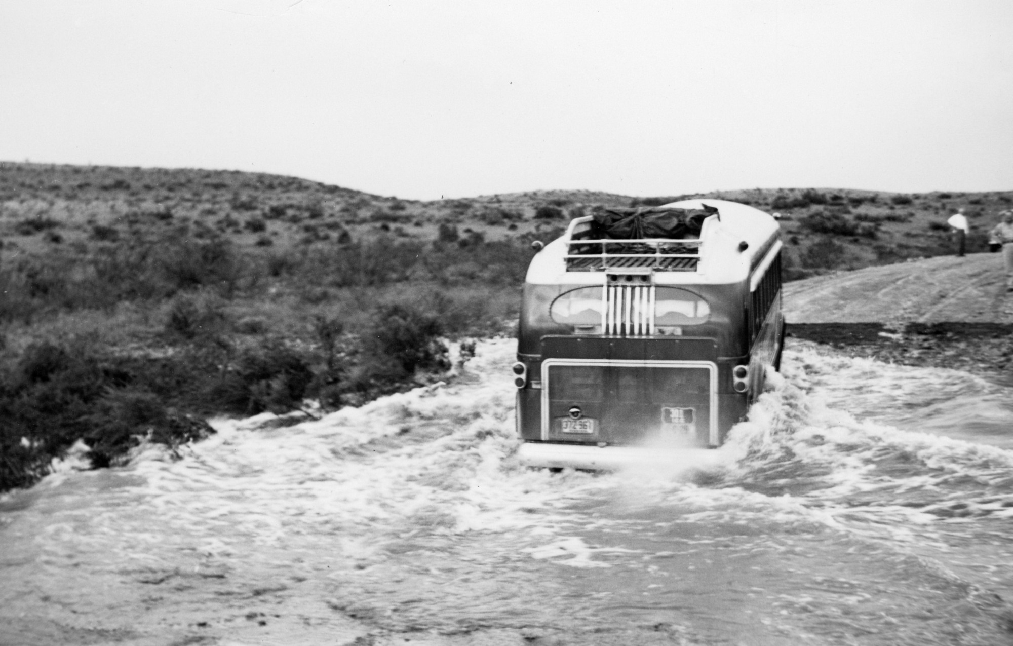 A black and white photograph of a bus attempting to drive through a partially flooded road.