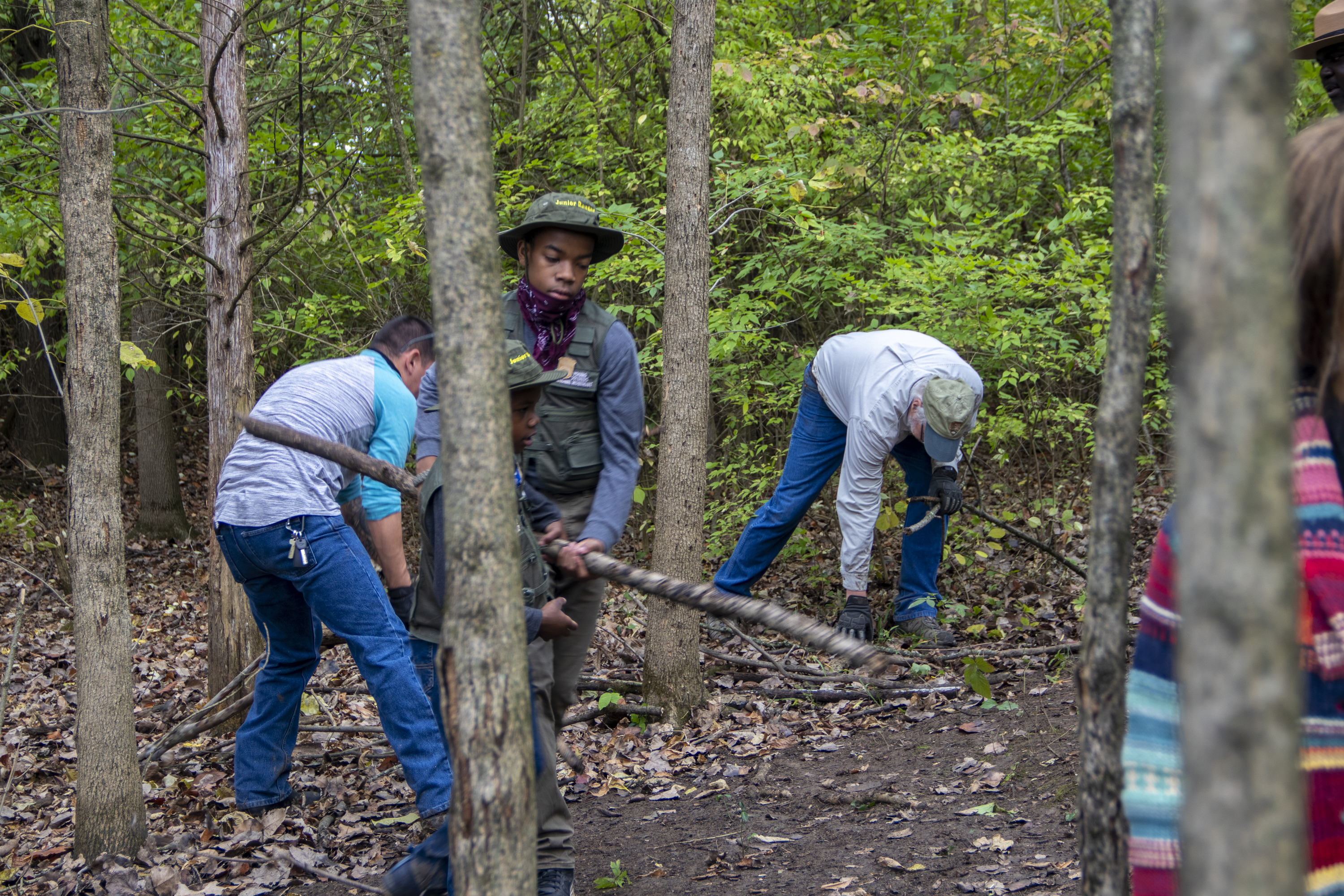 A young man in a green bucket hat carries a small tree limb from behind small trees.
