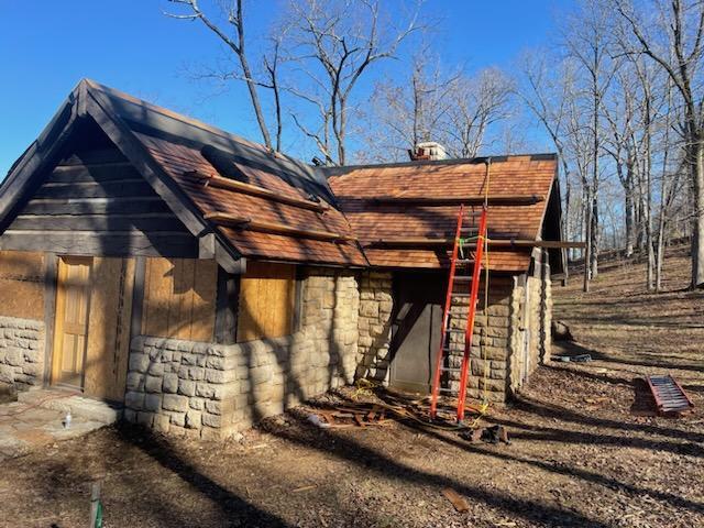 The same building as described previously, seen from the rear right, with new wood shingles nearly covering the roof. Foot Boards are attached to the roof and an orange ladder leans against the side of the building. Cloudless blue sky is visible in the background beyond the roof.