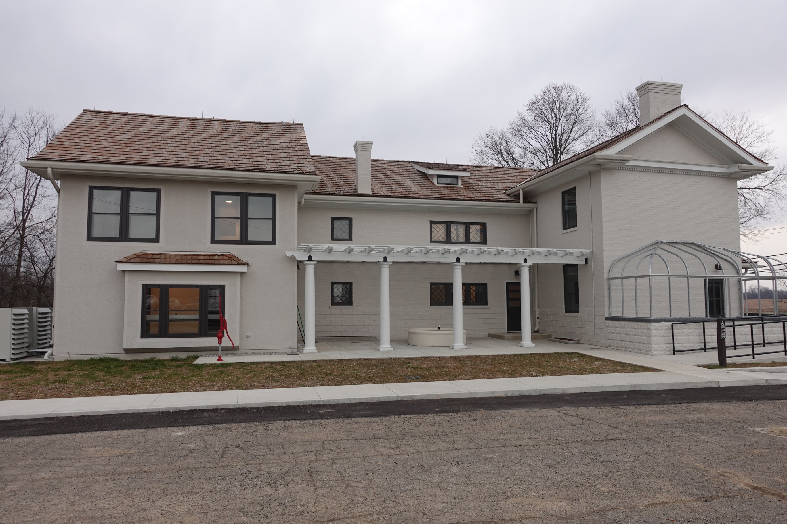 Grey two-story brick home with a red pump on the left and circular fountain under a pergola. 