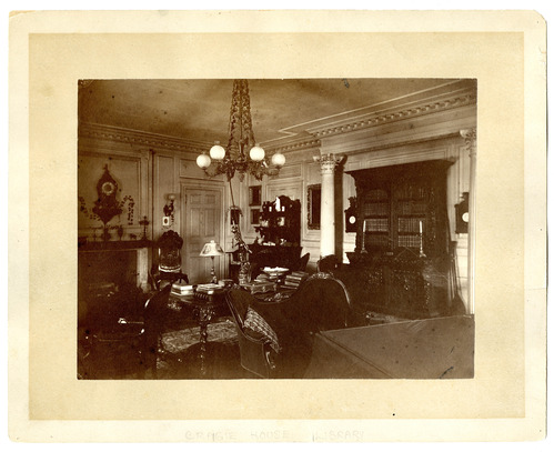 Black and white photograph of large 19th century library, crowded with dark furniture.