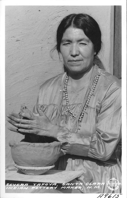 Black and white photo of woman staring at the camera while rubbing clay between her hands for the partially completed bowl in front of her.