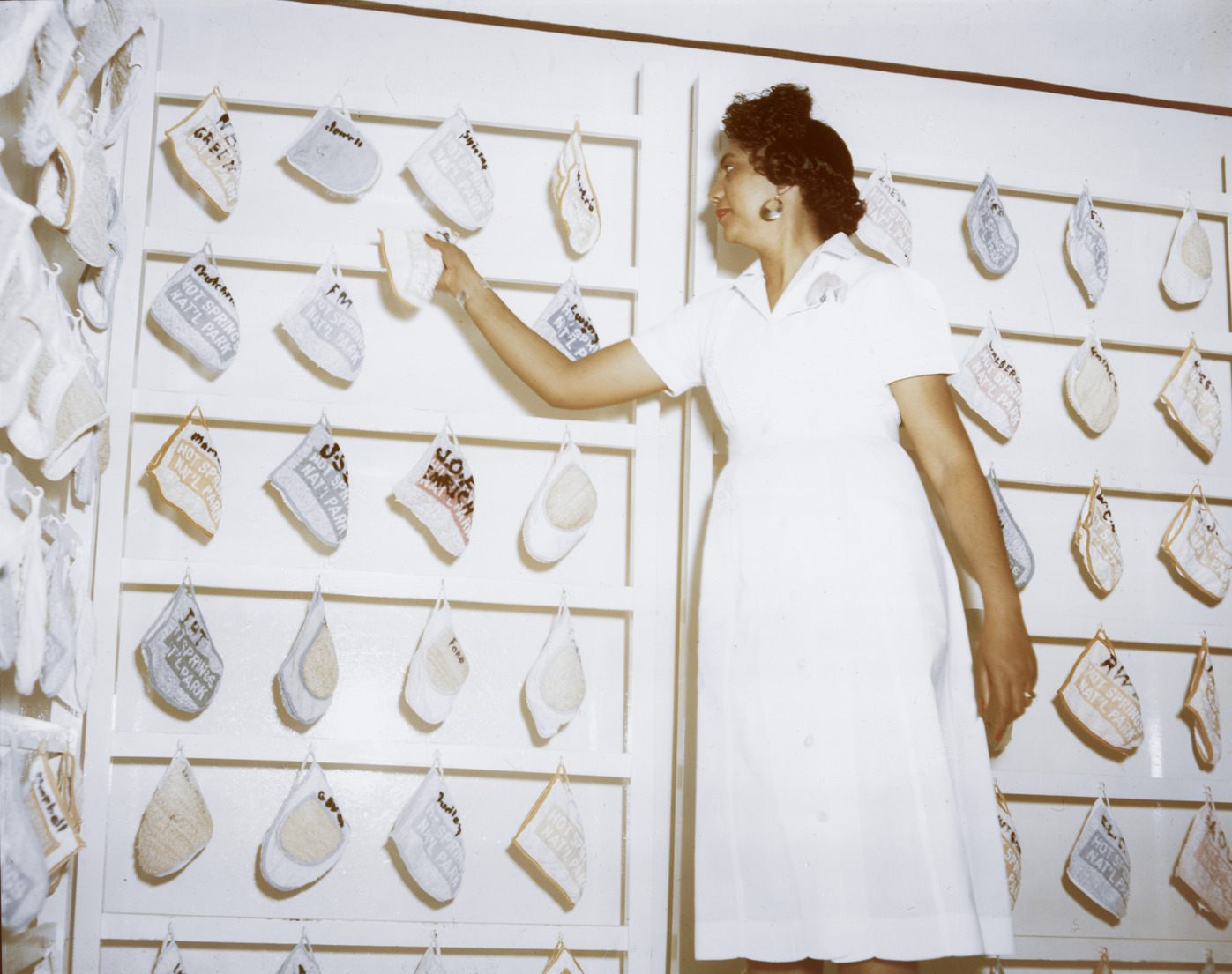 An African American woman in an all-white uniform stands in front of a wall lined with five rows of different colored mittens. She is reaching for one of the mittens with her right hand. 