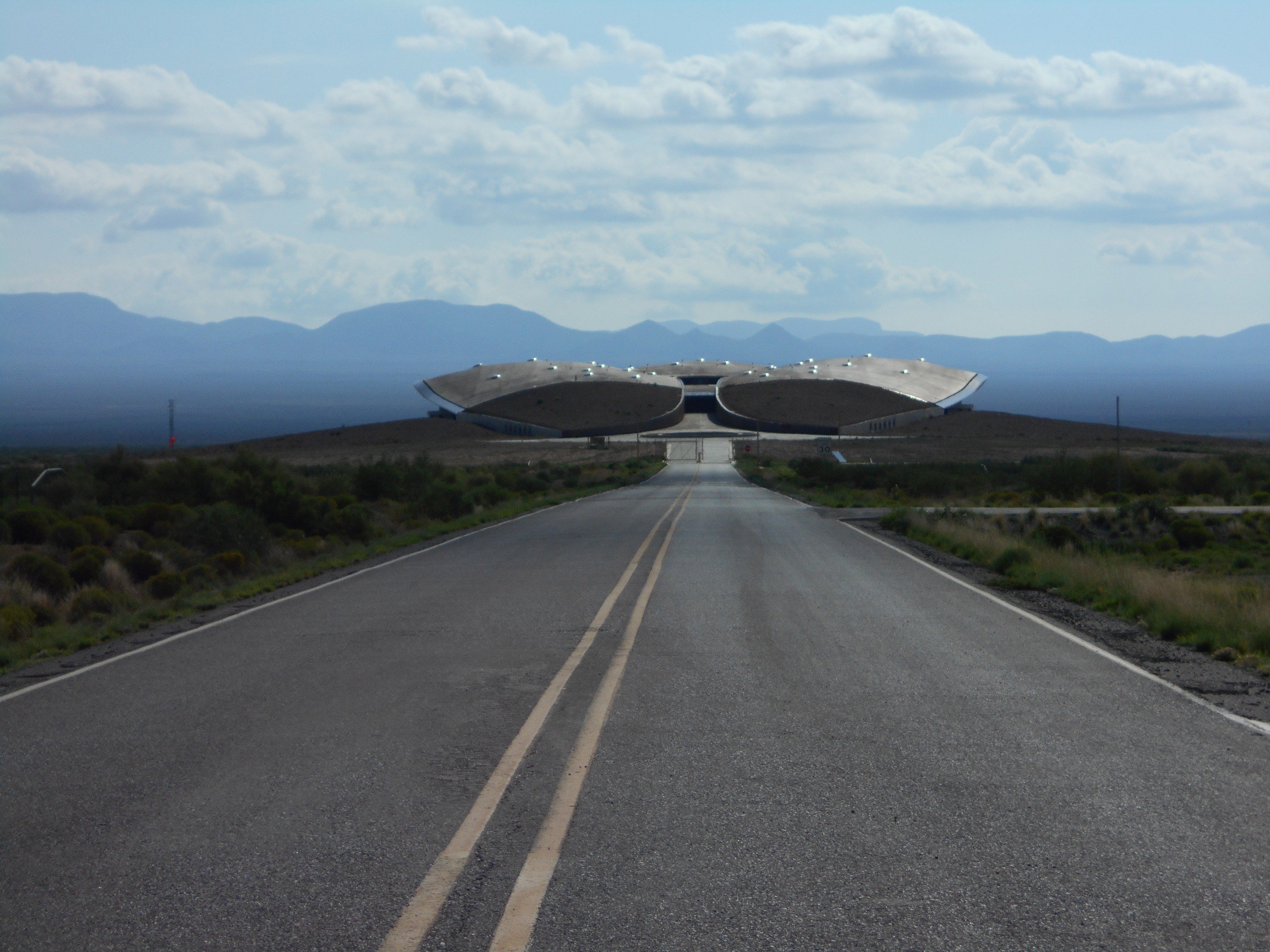 A straight road leading towards unique, futuristic buildings with mountainous backdrop under a cloudy sky.