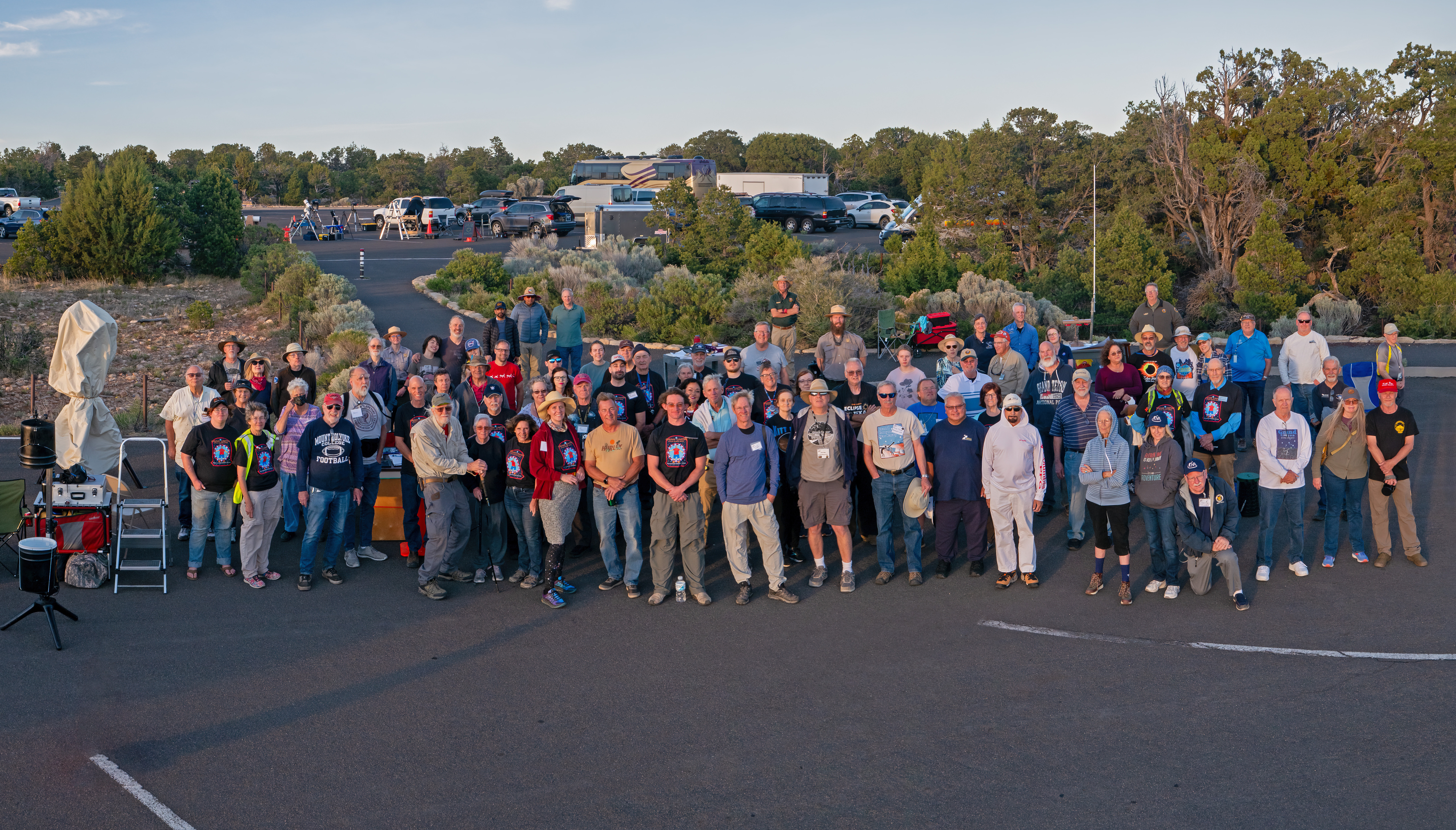 Just after sunset. a group of around 70 people are posing for a group photo on the edge of a large parking lot adjoining a forested area.