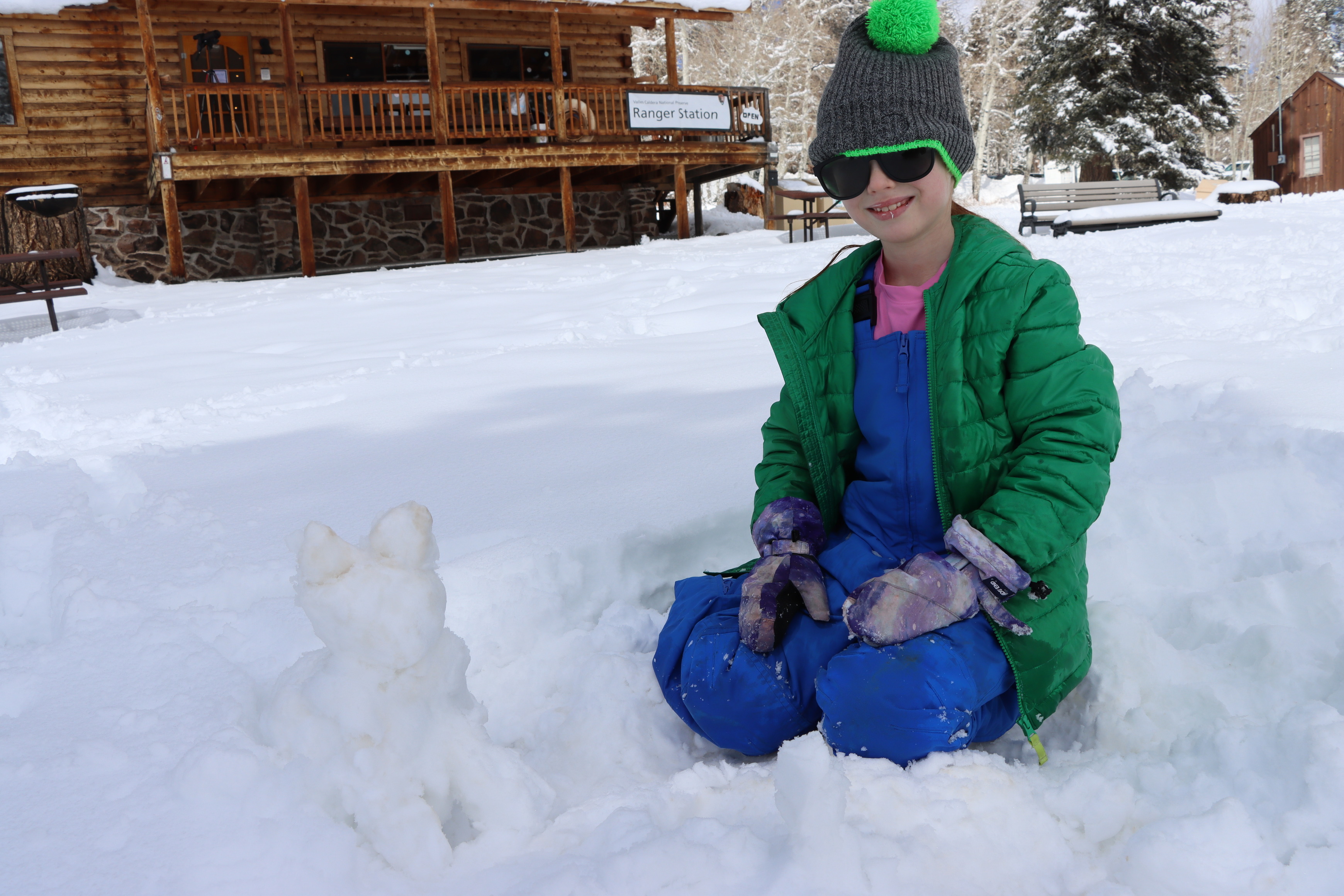 A child poses next to a cat-shaped snow sculpture in front of a log building.