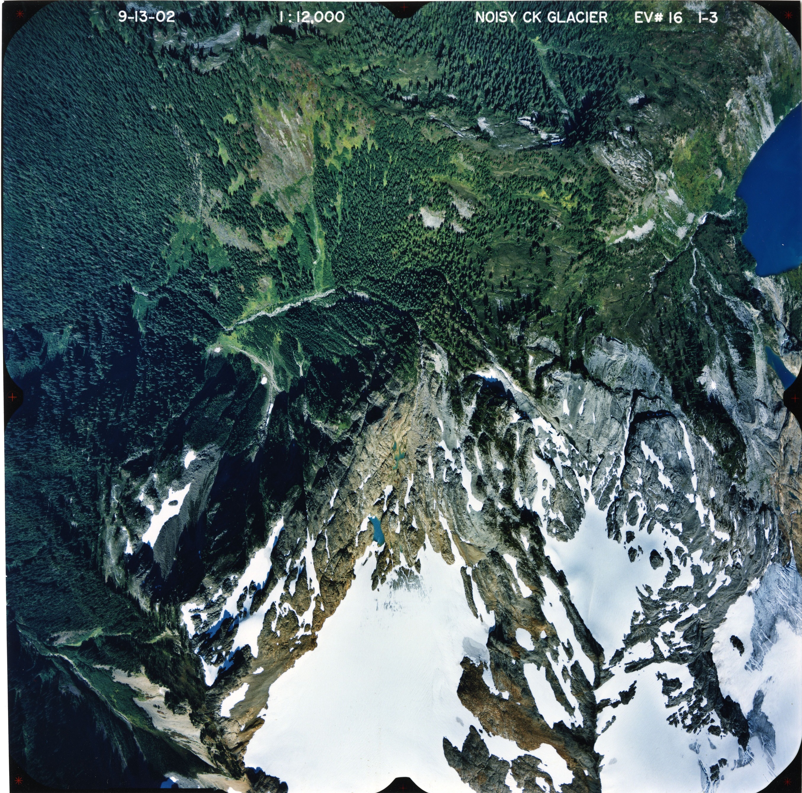 Color aerial photograph of glacier and alpine lake on mountain peak, surrounded by forested valleys.