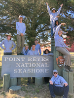 Ten teens stand around or sit on a gray wooden sign that says 