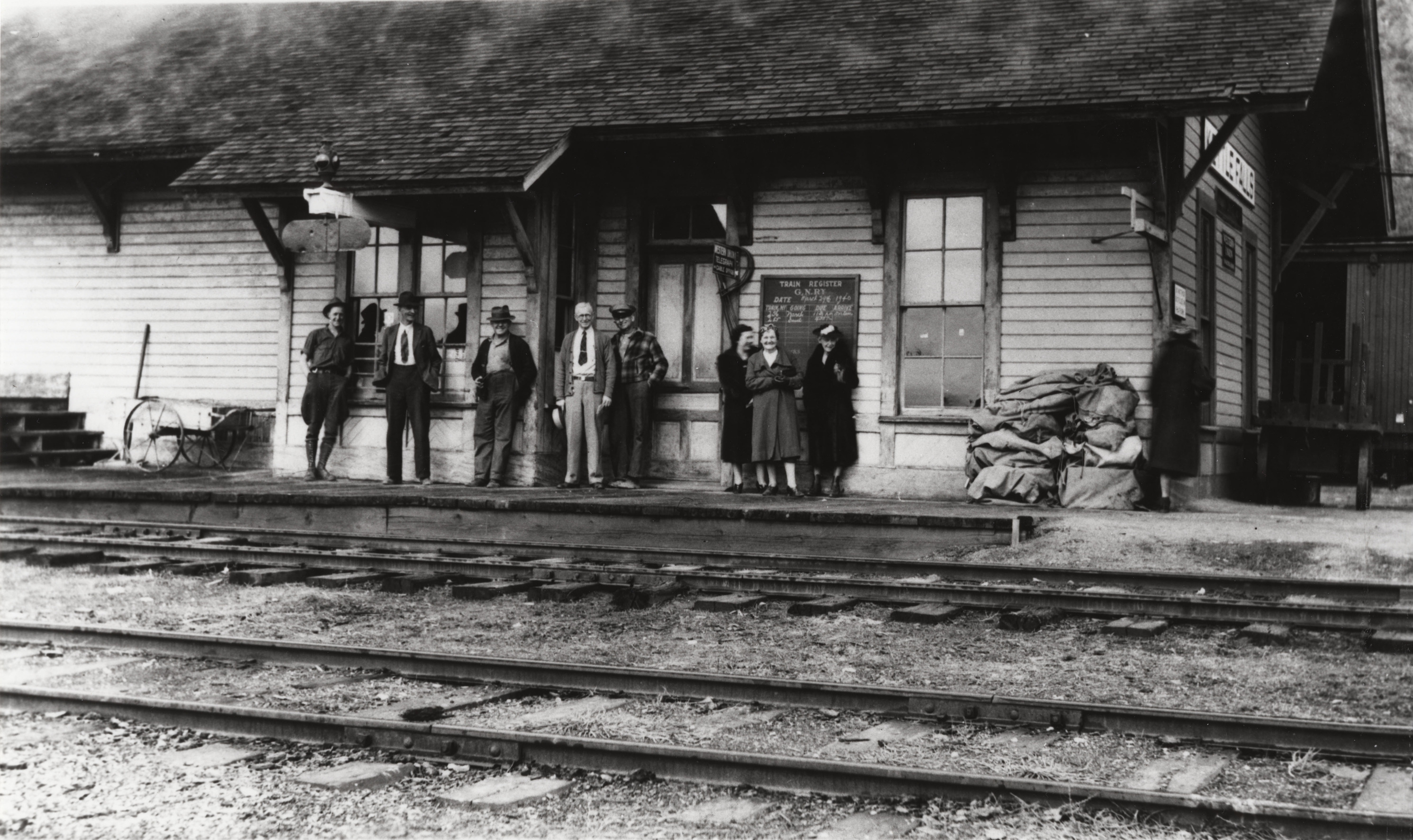 Black and white photo of a line of people waiting at a railroad station. 