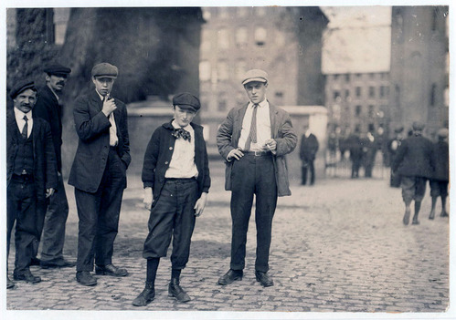 Five male workers in flat caps stare at the camera, some are child laborers. Other workers are in the background on the cobblestone path.