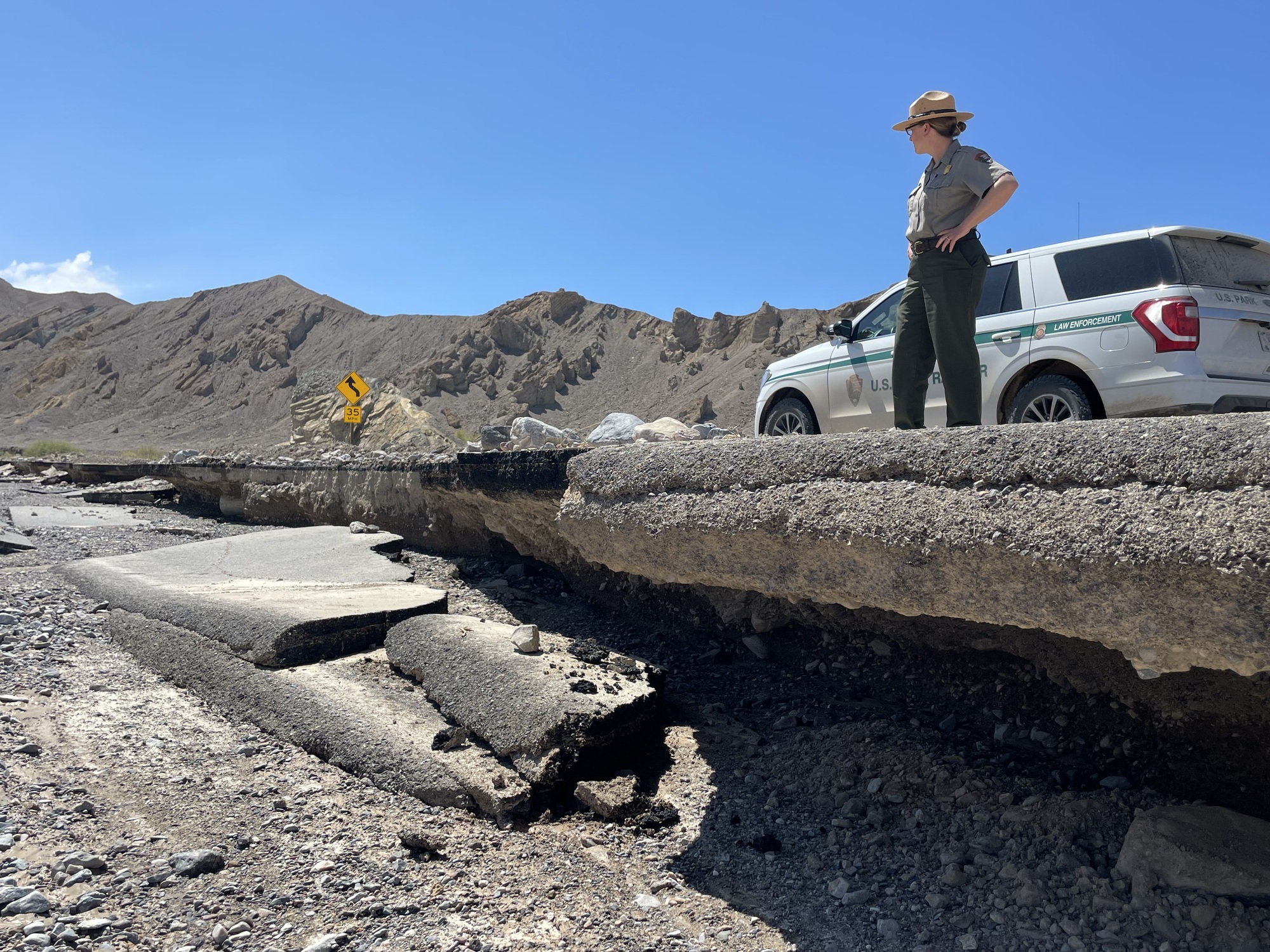 Side view of an undercut road with pavement which has fallen into the wash below and a ranger standing above on solid ground with a ranger vehicle and brown desert hills in the background.