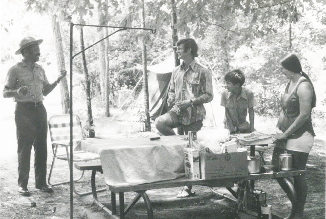 An African American man in a Ranger uniform talks to a smiling white family at a picnic table in the woods. 