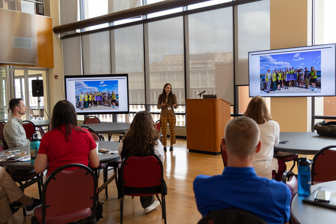 A woman presents in the front of a group of people in a large room full of windows. 