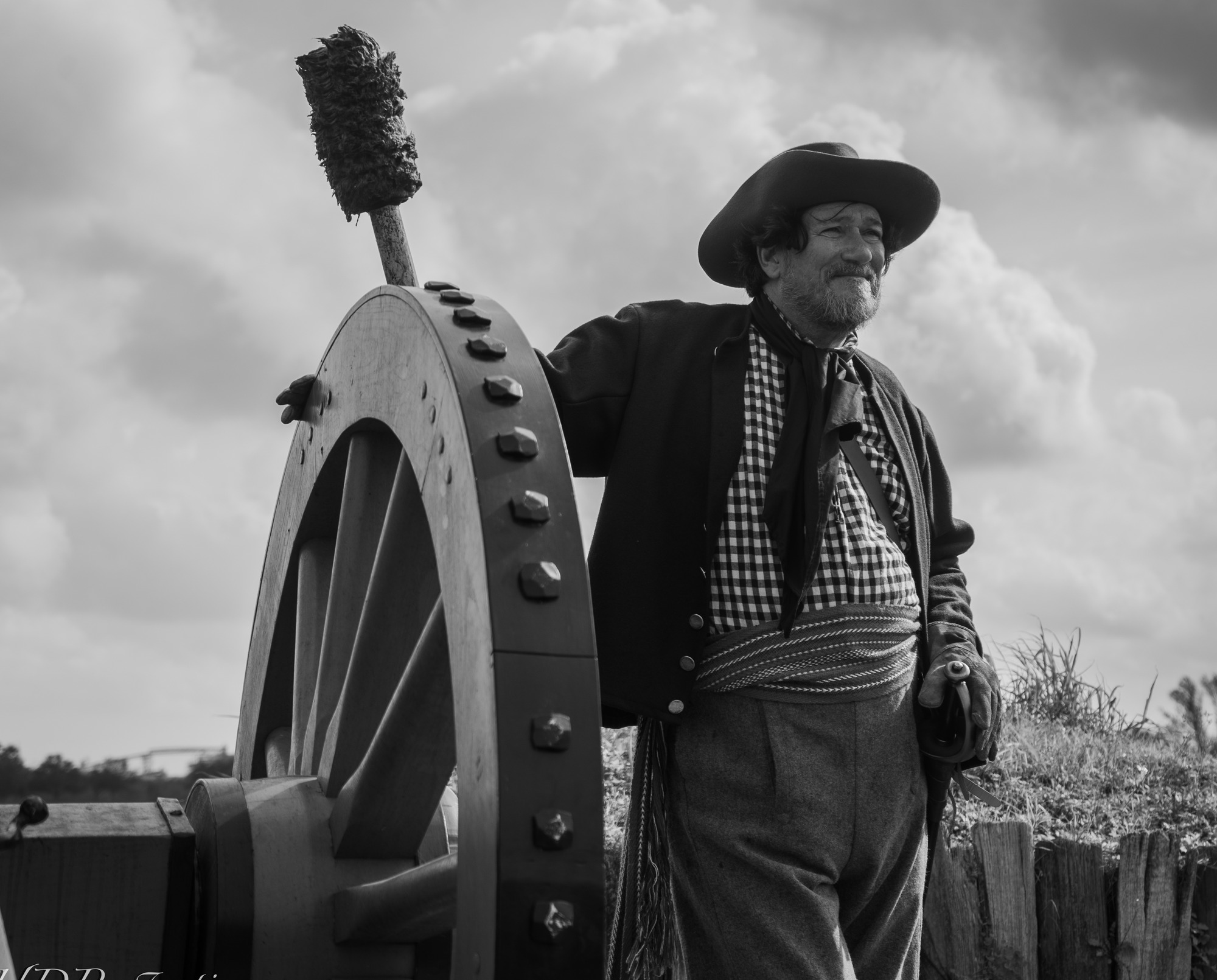 Black and white picture. Outside on a field. Man in period clothing stands next to a cannon wheel.