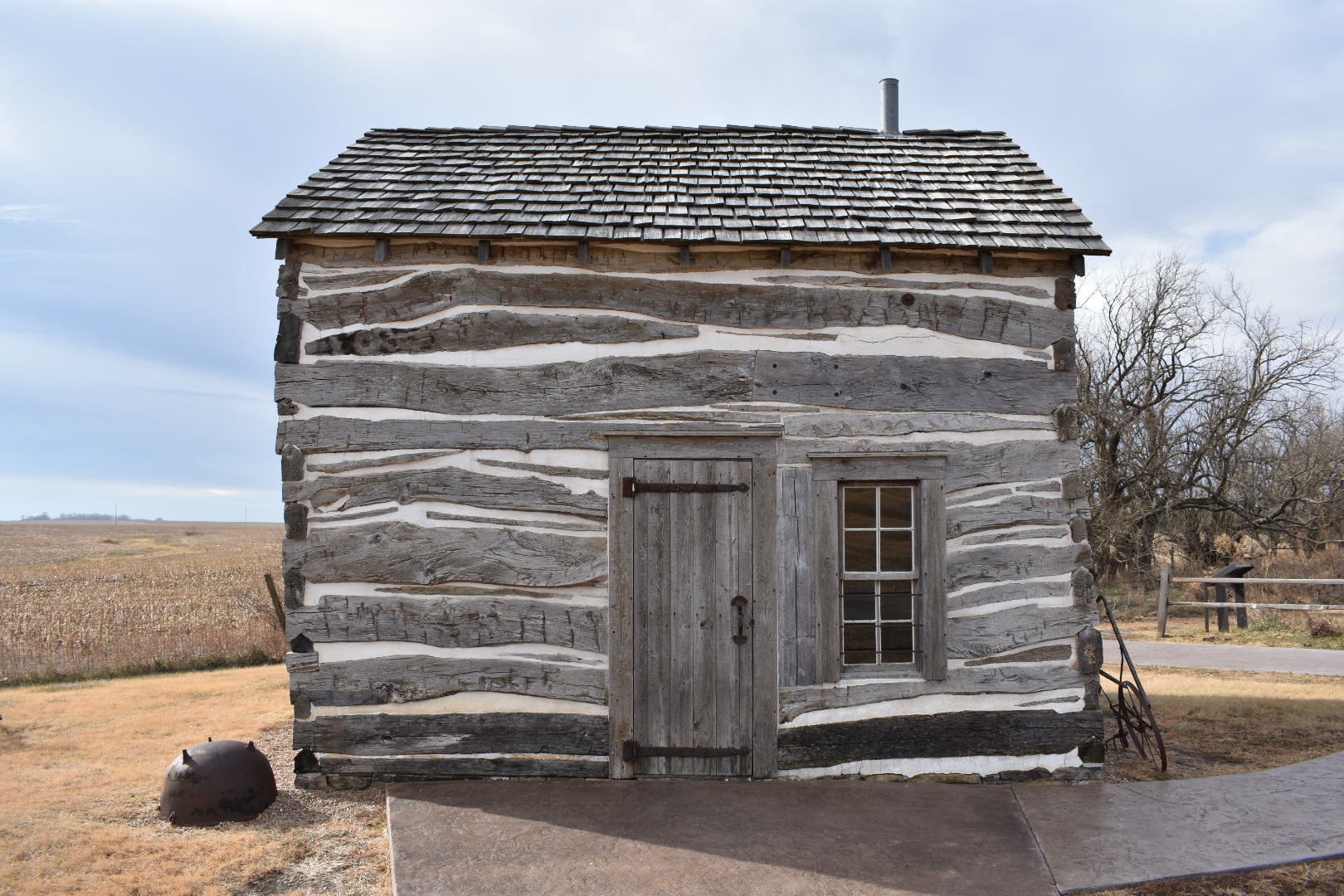 A one-and-a-half story log cabin has a shingle roof and a wooden door and single window in one side.