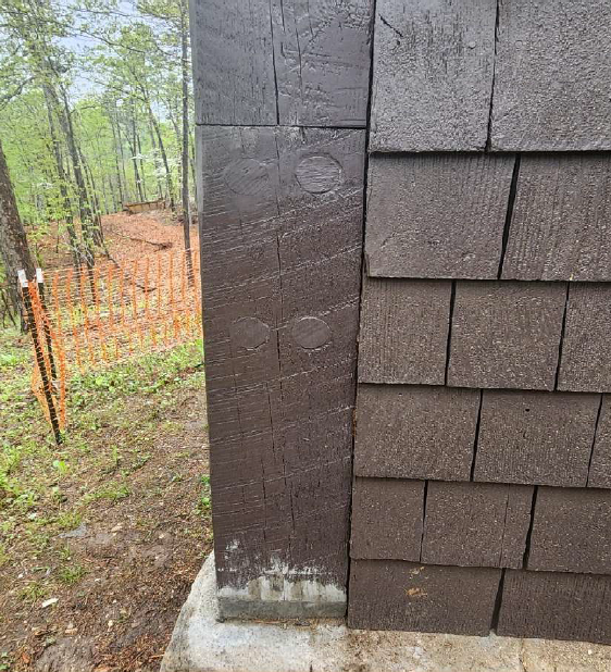 A wooden beam attached to the side of a brown painted, shingle sided building is centered in the frame. The building is on the right, and green forest with an orange mesh construction barrier in the foreground can be seen to the left. The wooden beam is painted brown like the building and has a dividing line where two sections have been attached. Four round holes in a rectangular pattern have been filled with wooden plugs and painted over.
