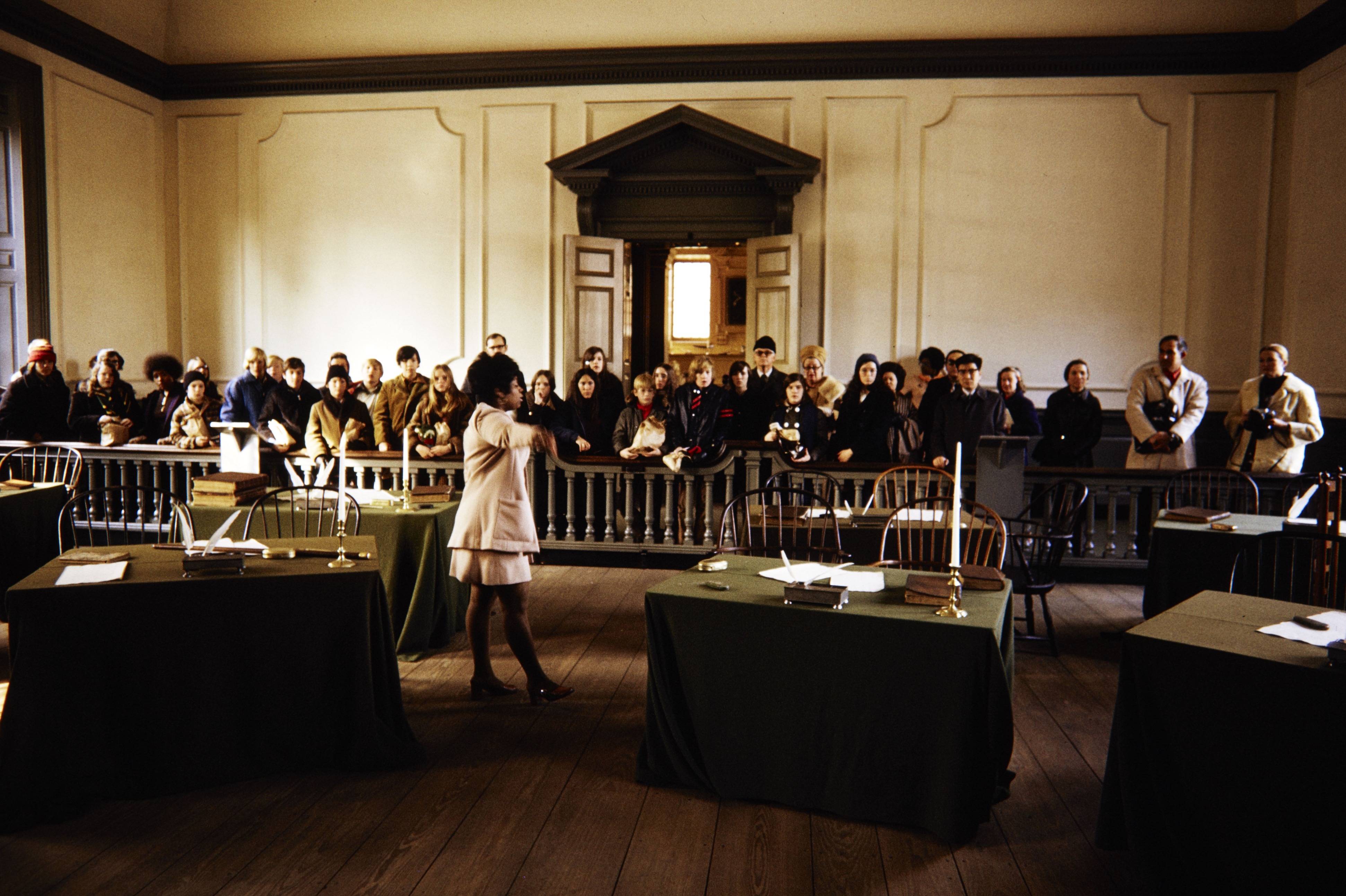 Woman in NPS uniform stands between two tables, addressing a large group of visitors standing behind a railing.