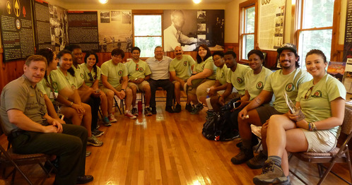 a park ranger and a group of about 15 people sit indoors in chairs in a horseshoe shape and smile at the camera. 