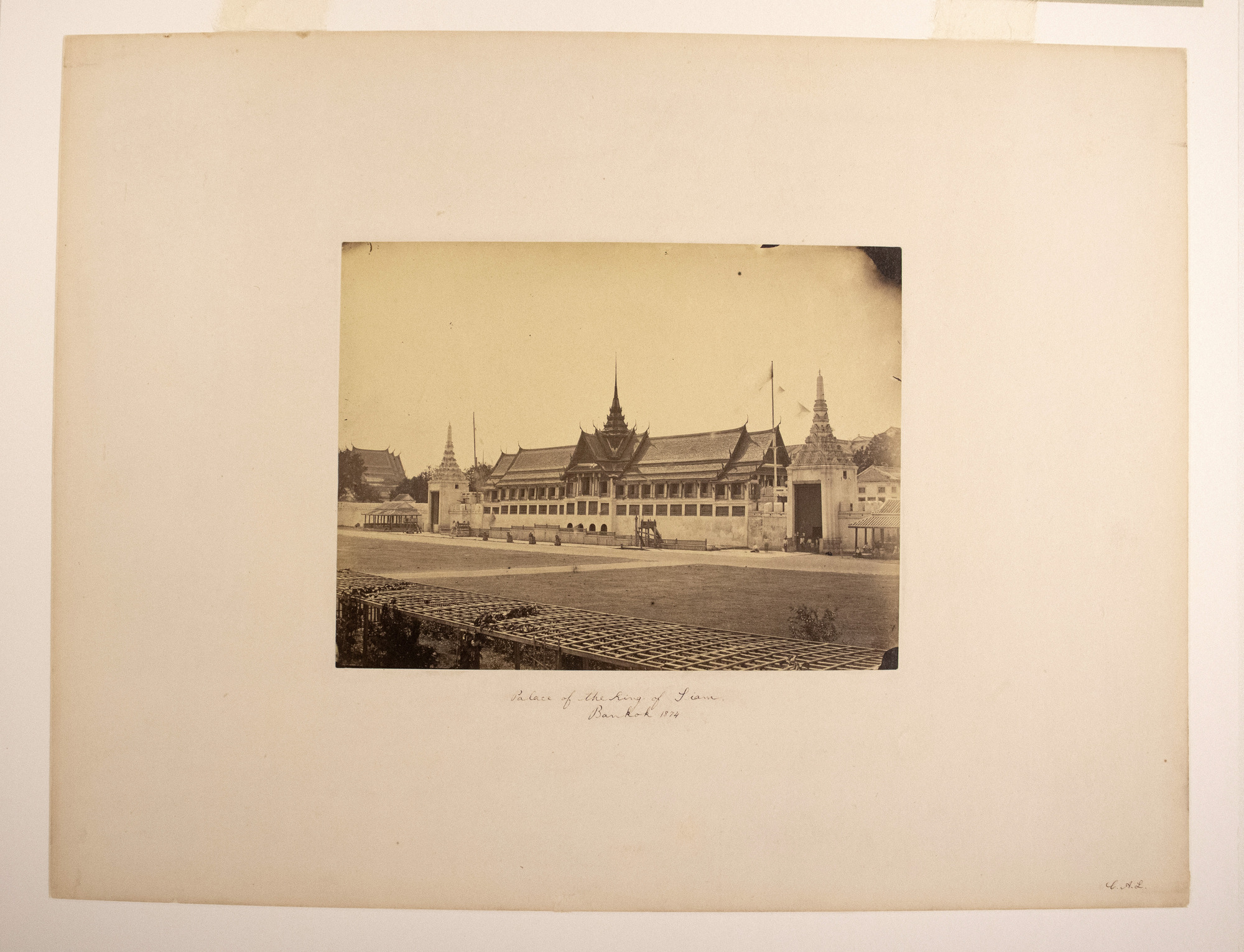 Sepia photograph with an inscription representing a long building with pointed parts of a roof, a few people standing by its wall, a road and lattice panels in the foreground.